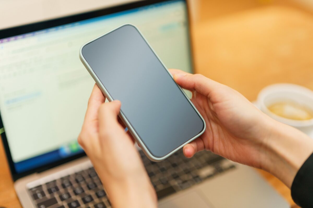 Ruky držia mobilný telefón nad notebookom, vedľa je šálka kávy.